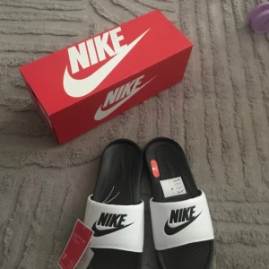  Nya sliders från Nike - Snygga och helt nya slidersfrån Nike i svart och vitt med stor logga över foten. Skorna har en bred rem i syntetmaterial och en räfflad sula för extra grepp. Perfekta att glida runt i på sommaren eller efter träningen.                  Funkar även för Size 41 och 42