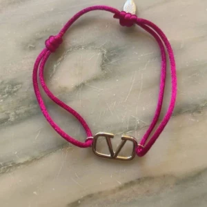 Valentino armband med rosa snöre - Snyggt armband från Valentino med ikonisk V-logga i metall. Armbandet har ett  rosa snöre med justerbara knutar och en liten metallbricka med Valentino-gravyr. Perfekt accessoar för att lyfta din outfit med en poppig färg och exklusiv känsla.