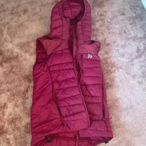 Mörkröd dunväst med huva 157 - Säljer en mörkröd dunväst från 157 i storlek S. Västen har en quiltad design, dragkedja framtill, praktiska fickor och en stor huva. Perfekt för lager-på-lager och chillare dagar. Justerbar nedtill med snören för bästa passform.