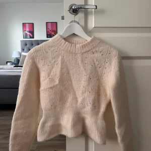 Ljusbeige stickad tröja med struktur - Supermjuk och fluffig ljusbeige stickad tröja med lång ärm och hög ribbad hals. Tröjan har ett diskret hålmönster och en lätt croppad passform som ger en snygg siluett. Perfekt för lager-på-lager under kyliga dagar.