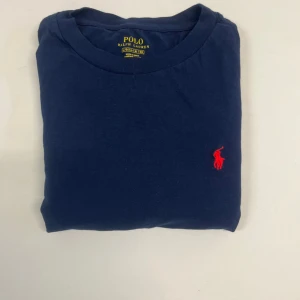  långärmad tröja från Polo Ralph Lauren - Snygg mörkblå långärmad tröja från Polo Ralph Lauren med klassisk röd broderad logga på bröstet. Tröjan är i mjuk bomull och har rund halsringning. Perfekt för en clean och stilren look. Har ett litet hål på axeln. Storlek 160