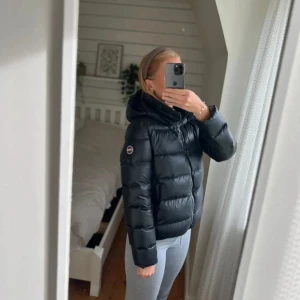 Svart colmar dunjacka - Säljer en svart pufferjacka med huva och dragkedja framtill. Jackan har en quiltad look och en badge på ärmen. Perfekt för kalla dagar och riktigt skön att bära. Passformen är normal och jackan är lätt att matcha med olika outfits. storlek 160 men passar en S/XS, har en liten skavank på vänster arm men inget som syns alls.