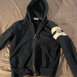 Mörkblå Moncler hoodie med logga - Snygg mörkblå hoodie från Moncler med dragkedja och huva. Tröjan har vita ränder och broderad Moncler-logga på ena ärmen. Köpt för 2000kr på kaspersheat, använd 2 gånger men den är liiite liten så därför säljer jag den