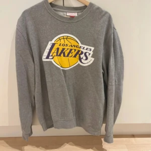 Grå Lakers sweatshirt i bomull - Säljer en grå sweatshirt med stort Los Angeles Lakers-tryck på bröstet. Tröjan har rund hals, långa ärmar och är gjord i mjuk bomull. Perfekt för dig som gillar basket och vill ha en chill och sportig stil.
