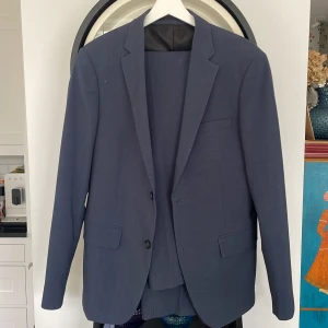 Blå kostym Lindbergh White slim fit - Snygg blå kostym från Lindbergh White i slim fit-modell. Kavajen har klassiska slag, två knappar och fickor med lock. Kostymen är tillverkad i polyester och har en stilren, modern look som passar perfekt när du vill vara extra uppklädd. Storlek 48 på kavajen och 46 på byxorna