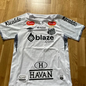 Santos Neymar Jr. 10 matchtröja Umbro - Vit Santos FC fotbollströja med Neymar Jr. och nummer 10 på ryggen. Tröjan har svarta tryck, flera sponsorer och klubbmärke på bröstet. Tillverkad i lätt och ventilerande material, perfekt för match eller träning. Klassisk passform med korta ärmar och subtilt mönster i tyget.