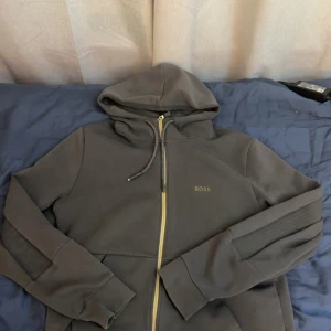 Mörkgrå hoodie från BOSS med dragkedja - Snygg mörkgrå hoodie från BOSS med guldfärgad dragkedja och huva. Tröjan har snörning i huvan, ribbade detaljer på ärmarna och diskret BOSS-logga på bröstet. Perfekt för en chill och stilren look.