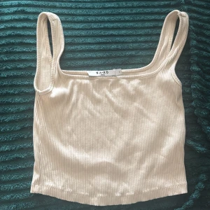 Beige ribbad linnetopp från NA-KD - Säljer en beige ribbad linnetopp från NA-KD. Toppen har breda axelband och är croppad med en enkel, stilren design. Perfekt för varma dagar och enkel att matcha med jeans eller kjol.