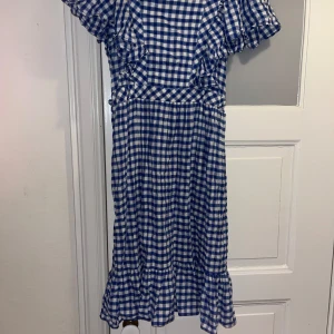 Blårutig kortklänning med volang - Supersöt kortklänning i blåvitt rutigt mönster med volangdetaljer vid ärmar och nederkant. Klänningen har v-ringning och korta ärmar, perfekt för sommarens alla tillfällen. Lätt och luftig i bomull, ger en fräsch och trendig look.