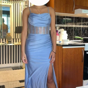 Ljusblå långklänning med strassdetaljer - Säljer en ljusblå långklänning med smala axelband och glittrande strassdetaljer över midjan. Klänningen har draperad överdel, figurnära passform och en hög slits på sidan. Materialet är mjukt och följsamt, perfekt för bal eller fest. Dragkedja i ryggen för enkel påklädning. OBS! använd en gång! köpte den för 4500kr
