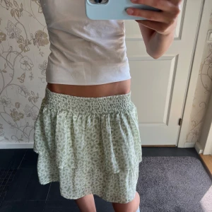 Grön blommig kortkjol från Kappahl - Supersöt kortkjol från Kappahl i ljusgrönt och vitt med blommigt mönster. Kjolen har bred smock i midjan och volangdetaljer som ger en flowy känsla. Tillverkad i mjuk bomull med innertyg för extra komfort. Perfekt för dig som gillar fräscha och somriga vibes!