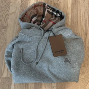 Grå Burberry hoodie  - Snygg grå hoodie från Burberry i storlek M med klassiskt rutigt foder i huvan och broderad logga på bröstet. Tröjan har snörning vid huvan, ribbade muddar och en stor magficka. Perfekt för dig som gillar stilrena designer med lyxig touch.