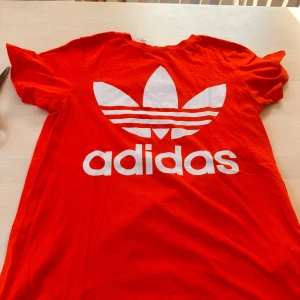 Röd Adidas t-shirt med logga - Snygg orange t-shirt från Adidas i storlek L med stor vit trefoil-logga och Adidas-tryck på bröstet. Klassisk rund hals och korta ärmar. Tillverkad i mjuk bomull som är skön mot huden. Perfekt för dig som gillar sportiga och stilrena plagg.