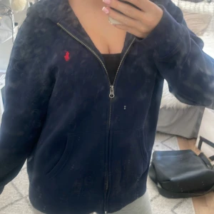 Mörkblå hoodie från Polo Ralph Lauren - Säljer en mörkblå hoodie från Polo Ralph Lauren med röd logga broderad på bröstet. Tröjan har dragkedja framtill, huva och två fickor. Materialet är mjukt och känns fleece-aktigt på insidan, perfekt för chill dagar. Passformen är loose och ärmarna är långa. Nypris: 999kr