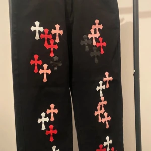 Svarta byxor med korsmönster från Shein - Svarta byxor från Shein i storlek XL med coola korsmönster i rött, vitt, rosa och svart längs benen. Materialet är en mix av bomull, polyester och viskos. Byxorna har raka ben och fickor bak, perfekt för dig som gillar statement-plagg.