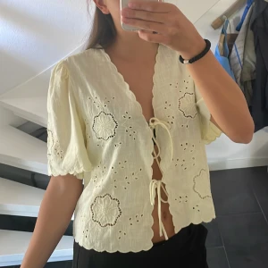 Ljusgul blus med broderi i storlek M - Superfin ljusgul blus från Gina Tricot i storlek M. Blusen har korta puffärmar, v-ringning och knyt framtill. Den är dekorerad med broderade blommor och spetsdetaljer. Blusen är i nyskick och har aldrig använts!