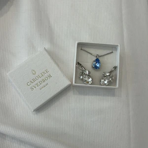 Caroline Svedbom smyckeset blå & silver - Snyggt smyckeset från Caroline Svedbom med ett halsband med blå droppformad kristall och matchande örhängen med stora, glittriga klara stenar. Nypris ca. 1400kr💗☀️Opps örhängena är slitna å lite svåra att stänga. 