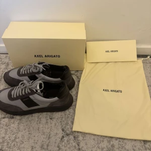 Axel Arigato skor - Snygga sneakers från Axel Arigato i grått och svart med stilren design. Skorna har ovandel i mocka och textil, svarta detaljer och platt sula. Klassisk snörning och logga på sidan. Perfekta för dig som gillar minimalistisk och modern stil. Skorna är använda ett fåtal gånger och är lite noppriga vid hälen men det är inget som stör och kan gissa på att de lätt kan tas bort. Lådan samt tillbehör följer med. Pris kan diskuteras vid snabb affär.