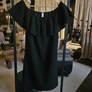 Svart offshoulder klänning med volang - Stilren svart kortklänning med offshoulder-modell och bred volang runt axlarna. Klänningen har en rak passform och är perfekt för dig som gillar en enkel men trendig look. Materialet känns mjukt och följsamt mot kroppen.