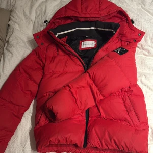 Röd pufferjacka från Calvin Klein Jeans - Riktigt snygg röd pufferjacka från Calvin Klein Jeans med huva och dragkedja. Jackan har svarta och vita detaljer på insidan samt logga på bröstet även på vänster arm och insidan. Perfekt för kalla dagar och har en cool, sportig vibe.