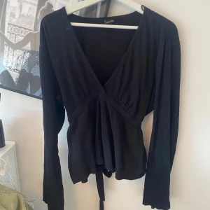 Svart topp från Gina Tricot XL - Svart och fin topp från Gina Tricot i storlek XL. Toppen har djup v-ringning, långa utsvängda ärmar och knytband i midjan som ger en snygg siluett. Materialet är mjukt och lätt strukturerat, perfekt för att lyfta din outfit med en enkel men stilren look.