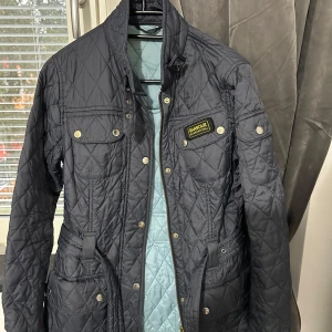 Quiltad svart jacka från Barbour - Säljer en svart quiltad jacka från Barbour International med snygga detaljer som bälte i midjan, tryckknappar och dragkedja. Jackan har flera fickor framtill och klassisk Barbour-logga på bröstet. Perfekt för dig som gillar stilren och sportig look.