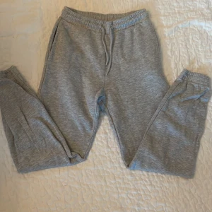 Grå mjukisbyxor från KappAhl, stl 158 - Mysiga grå mjukisbyxor från KappAhl med resår i midjan och benslut. Byxorna har dragsko i midjan och är tillverkade i mjukt bomullsmaterial. Perfekta för chill eller träning. Klassisk rak passform och neutralt utseende.