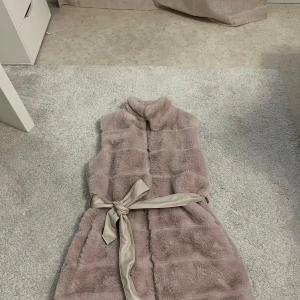 Beige pälsväst med bälte  - Supermjuk och fluffig beige pälsväst från new Yorker med hög krage och ett snyggt satinband i midjan som bälte. Västen är fodrad med glansigt tyg och har en stilren, rak passform som ger en lyxig vibe.