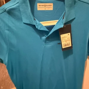 Blå pikétröja från Henri Lloyd - Snygg blå pikétröja från Henri Lloyd i klassisk modell med krage och knappar framtill. Tröjan är i bomull och har en stilren logga broderad på bröstet. Perfekt för dig som gillar en sportig och fräsch look.