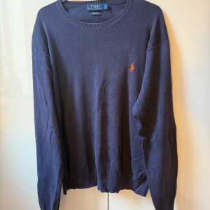 Mörkblå tröja från Polo Ralph Lauren - Klassisk mörkblå tröja från Polo Ralph Lauren med rund halsringning och den ikoniska broderade loggan i orange på bröstet. 