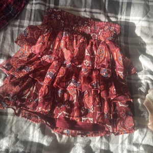 Röd volangkjol med paisleymönster - Supersöt kort kjol i rött med färgglatt paisleymönster och flera volanger. Resår i midjan för skön passform. Perfekt för dig som gillar boho-vibe och vill sticka ut lite extra.