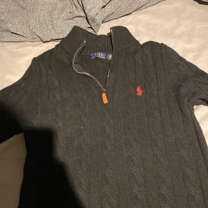Svart stickad half zip tröja Polo Ralph Lauren - Svart kabelstickad tröja från Polo Ralph Lauren med half zip och ribbad krage. Klassisk röd logga broderad på bröstet. Tröjan har långärmade och är gjord i mjukt stickat material, perfekt för kyliga dagar.