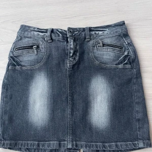 Mörkgrå jeanskjol från Lindex, storlek 36 - Mörkgrå kort jeanskjol från Lindex i storlek 36. Kjolen har slitna detaljer, framfickor med dragkedja och klassiska bakfickor. 