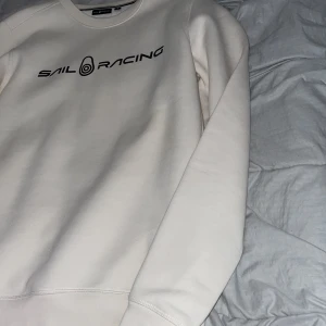 Vit Sail Racing sweatshirt i bomull - Fräsch vit sweatshirt från Sail Racing i storlek S. Tröjan har svart logotyptryck på bröstet och en liten logga i nacken. Rund hals och långa ärmar, tillverkad i mjuk bomull med ribbade muddar. Perfekt för chill dagar och enkel att matcha med jeans eller joggers.