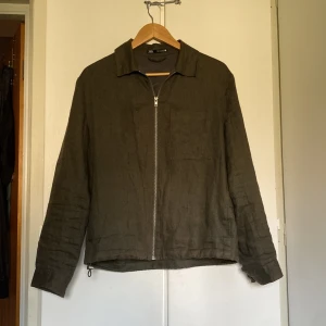 Olivgrön overshirt från Zara i linne - Snygg olivgrön overshirt från Zara i 100% linne. Jackan har klassisk krage, bröstficka och hel dragkedja framtill. Perfekt för lager-på-lager och chill stil. Passar dig som gillar minimalistiska och stilrena plagg.