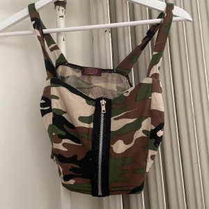 Camo korsett-topp  - Snygg korsett liknande topp med camouflage-mönster från Instyle. Toppen har breda axelband, dragkedja framtill och är croppad. Färgerna är grönt, brunt, beige och svart. 