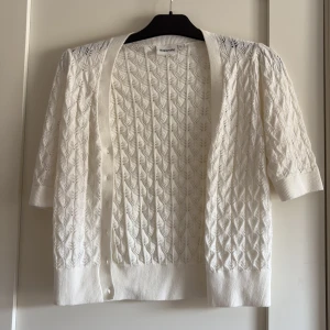 Vit stickad kofta från Kappahl - Säljer en söt vit stickad kofta från Kappahl i storlek S. Koftan har korta ärmar, fina knappar framtill och ett snyggt hålmönster som ger en fräsch och trendig look. Perfekt att slänga över en topp när det är lite kyligt ute.