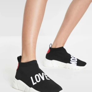 Love Moschino svarta sneakers - Svarta sneakers från Love Moschino med vit chunky sula och stickad ovandel. Skorna har texten 'LOVE MOSCHINO' i vitt och svart på ovandelen samt röd detalj vid hälen. Snygg strumpliknande passform och modern streetstil.