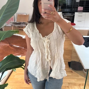 Romantisk vit blus med spets och knyt - Supersöt vit blus från hunkydorry med spetsdetaljer, knyt framtill och fina veck längs hela framsidan. Blusen har korta ärmar och en lätt, luftig känsla. Romantisk/bohemisk.