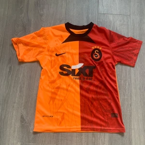 Galatasaray fotbollströja - Galatasaray fotbollströja från Nike med Dri-FIT ADV-material. Tröjan har korta ärmar, är delad i orange och röd med geometriskt mönster och klubbmärke på bröstet. Svart Nike-logga och sponsortryck på framsidan. Perfekt för dig som älskar fotboll och vill sticka ut. Skriv för frågor eller funderingar priset är inte huget i sten