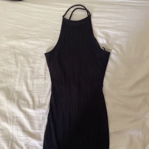 Svart ribbad kortklänning med snörning - Säljer en svart ribbad klänning med tight passform och snygg snörning i ryggen. Klänningen är ärmlös och har en hög hals med tunna axelband. Perfekt för dig som gillar minimalistisk stil och vill ha något som sticker ut med detaljer i ryggen. Använd endast en gång!