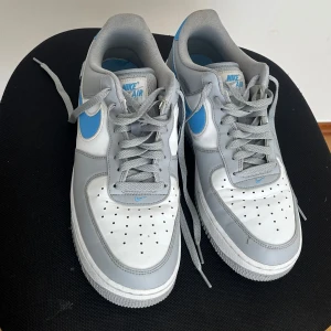 Nike air force 1 i grått och blått  - Nike Air Force 1 sneakers i en clean färgkombo av grått, vitt och blått. Klassisk låg modell med perforerad tå, grå snörning och blå swoosh. Ovandel i skinn och syntet, vit sula med AIR-detalj. Perfekta för dig som gillar streetstyle och vill sticka ut.
