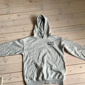 Grå hoodie från Gina Tricot med tryck - Väldigt bra skick och har använt den kanske 7+ gånger💗Grå hoodie från Gina Tricot med svart texttryck 'JE M'EN AMOUR' och en stjärna på bröstet. Mjuk insida, huva och ribbade muddar. Perfekt för chill dagar och enkel att matcha med jeans eller leggings.