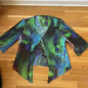 Färgstark overshirt från Elinette - Säljer en tunn och flowy blus från Elinette med galet snyggt mönster i blått, grönt, lila och svart. Jackan har trekvartsärmar, v-ringning och en liten metallknapp framtill. Perfekt för att lyfta vilken outfit som helst.