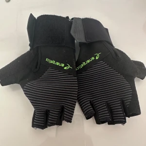 Svarta träningshandskar från Energetics - Säljer ett par svarta fingerlösa träningshandskar från Energetics med gråa ränder och grönt logotryck. Handskarna har kardborreband vid handleden och är tillverkade i syntetmaterial med perforerade detaljer för extra ventilation. Perfekta för gymmet eller cykling.