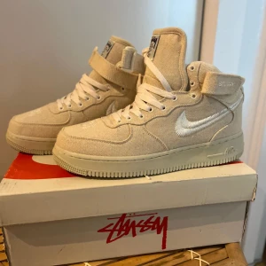 Nike Air Force 1 High Stussy beige - Nike Air Force 1 High Stussy i beige canvas med vita broderade swooshes och Stussy-detaljer på remmen och hälen. Klassisk high-top siluett med snörning och kardborreband. Platt sula och rund tå, perfekt för dig som gillar streetwear och unika samarbeten.