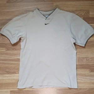 Vintage 1990s Nike t-shirt - Stilren 1990s vintage beige t-shirt från Nike med v-ringad hals och svart broderad swoosh på bröstet. T-shirten har korta ärmar och diskreta svarta detaljer vid hals och ärmslut. Perfekt för en clean och sportig look.