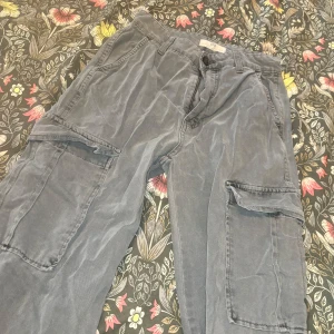 Grå cargobyxor med stora fickor - Snygga grå cargobyxor från STR med stora sidofickor och klassisk passform. Byxorna har bälteshällor, knapp och dragkedja framtill. Perfekta för en chill och avslappnad stil. Materialet känns mjukt och slitstarkt.