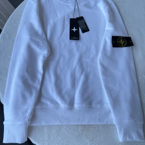 Vit Stone Island sweatshirt i bomull - Vit sweatshirt från Stone Island med klassisk rund hals och ikonisk logotyp-patch på vänster ärm. Tröjan är i mjuk bomull och har ribbade muddar vid ärmslut och nederkant. Perfekt för dig som gillar clean och stilren streetwear.