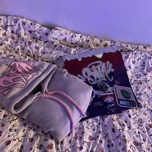 Grå hoodie från Unlucky Rules - Säljer en grå hoodie från Unlucky Rules med fet rosa graffiti-tryck på huvan och grova rosa snören. Klassisk känguruficka framtill och mjukt material som känns riktigt skönt. Perfekt för dig som gillar streetwear och vill sticka ut lite extra.